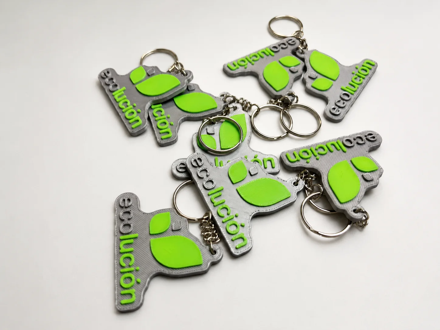 Llaveros personalizados impresos en 3D con el logo de una empresa en relieve, gris y verde, con la anilla montada