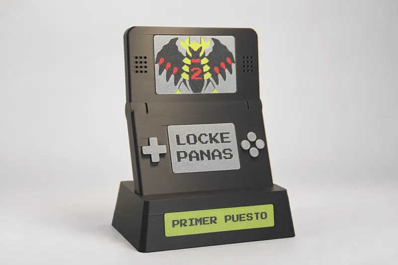 Trofeo impreso en 3D con forma de consola portátil abierta, en cuatro colores, con placa «PRIMER PUESTO» en la peana